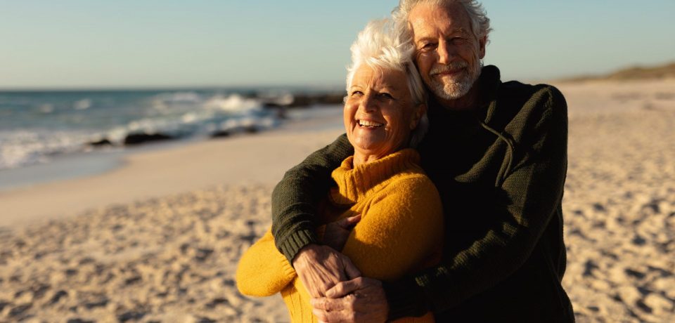 Front view of a senior Caucasian couple at the beach wearing sweaters, embracing and smiling to camera, with blue sky and sea in the background